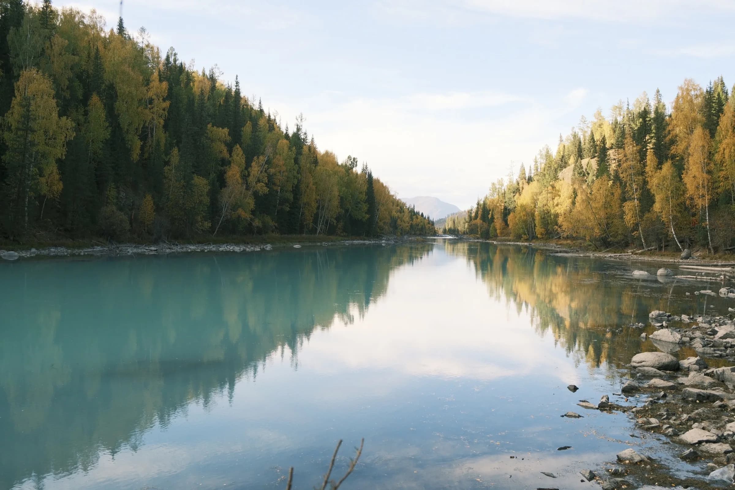 A still turquoise river reflecting autumn trees and distant mountains