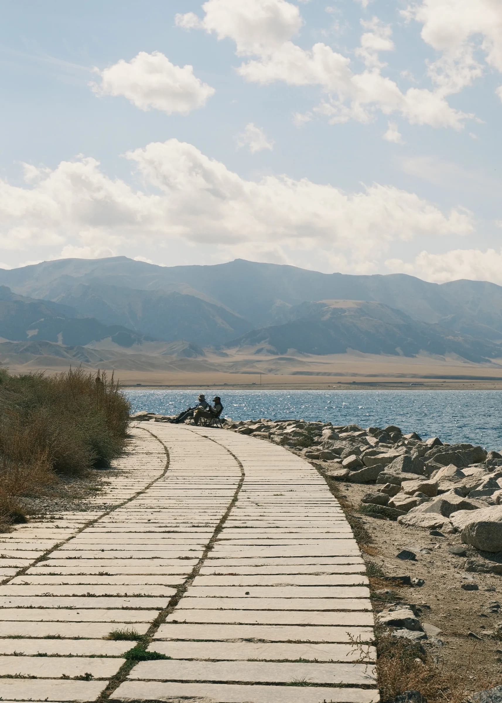 A curving stone path along the lakeshore toward two people sitting by the water